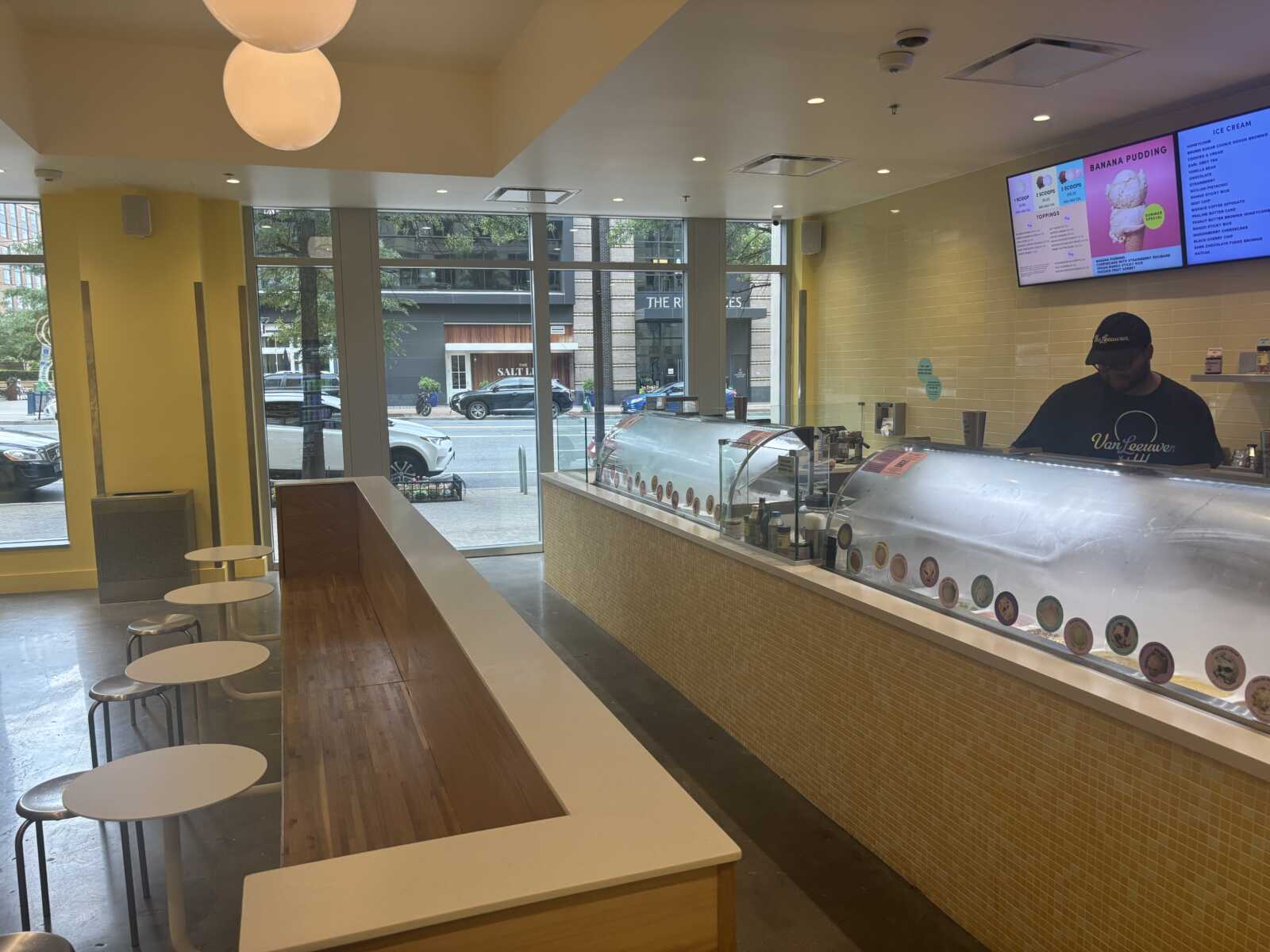 Bright ice cream shop interior with a long glass display case, yellow tiled wall, and seating along a white and wood counter; staff member behind the counter.