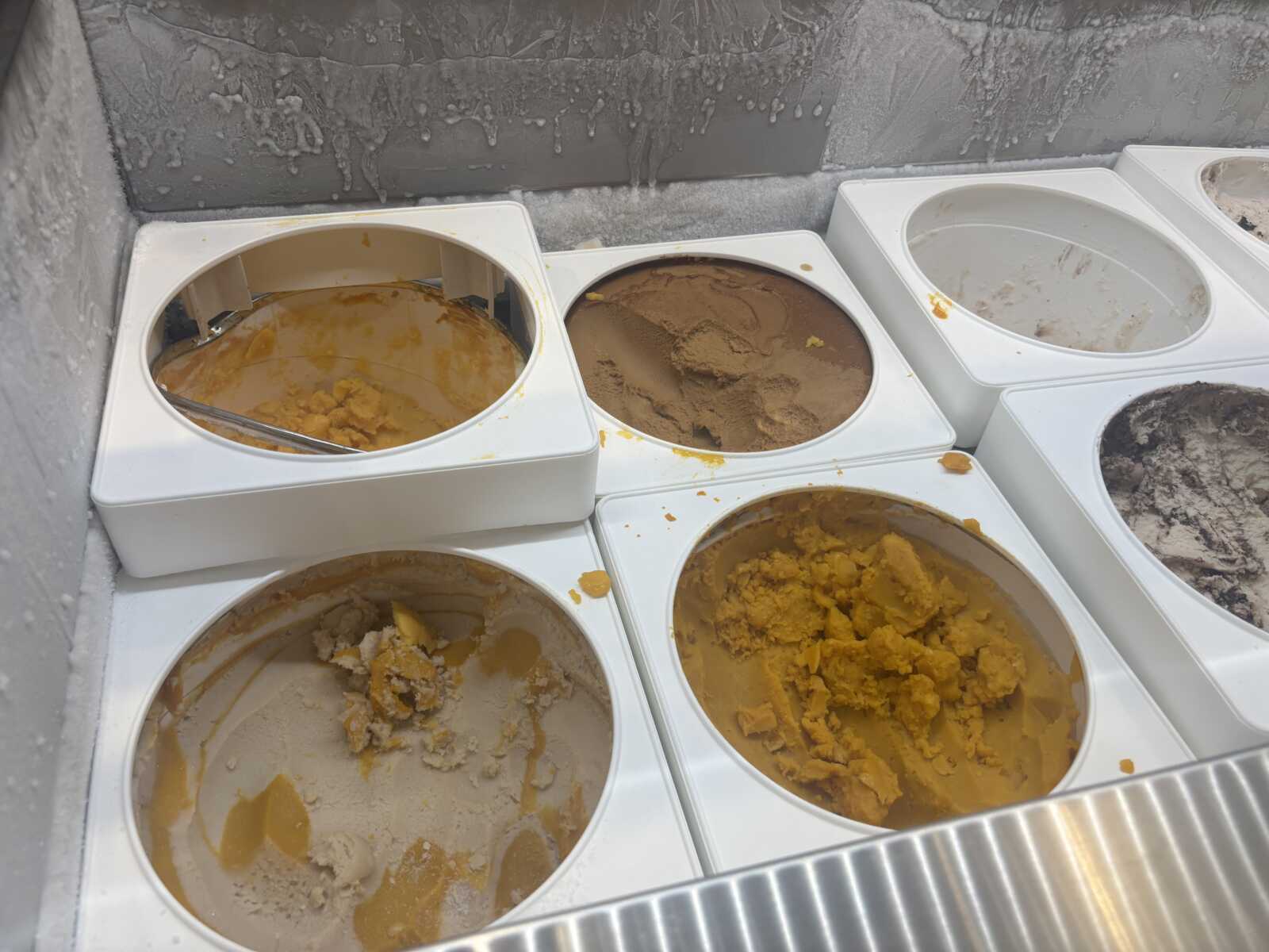 Open ice cream display with several metal tubs of colorful gelato flavors in yellow and brown hues.