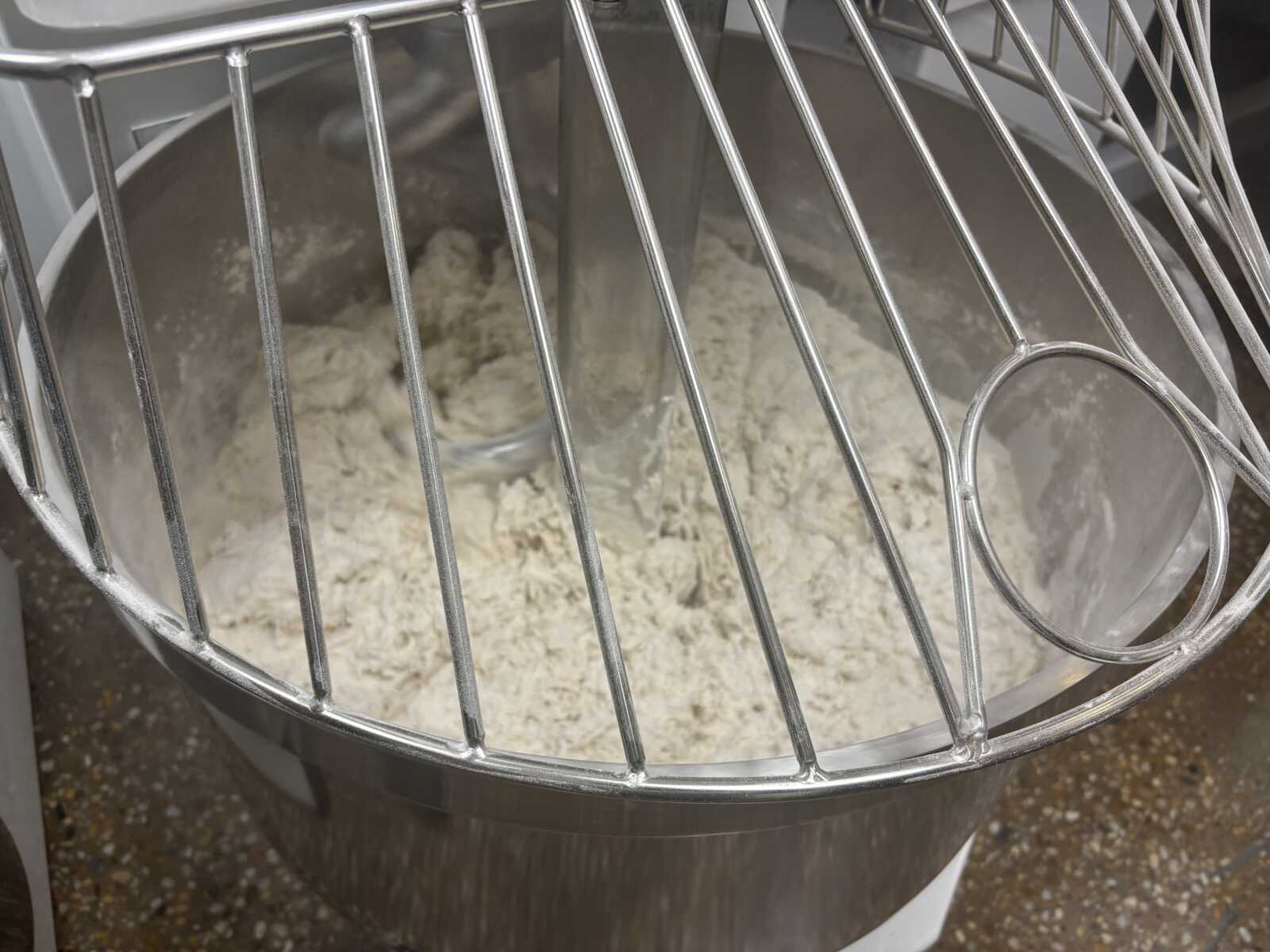 Close-up of a stainless steel stand mixer bowl with doughy flour inside and wire beaters visible in the foreground.