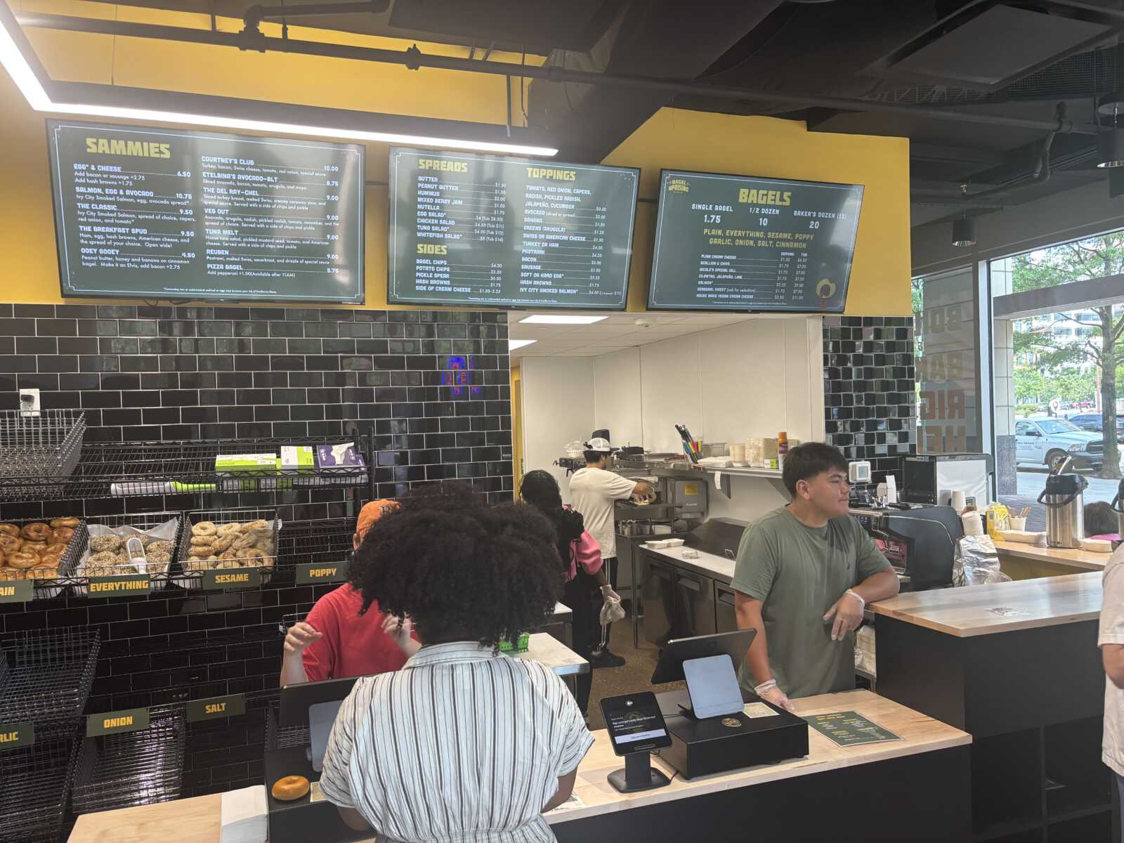 Inside a bagel shop with customers at the counter, three menu boards overhead, and baskets of bagels (onion, sesame, poppy, salt) on a black-tiled rack fronting the wall.