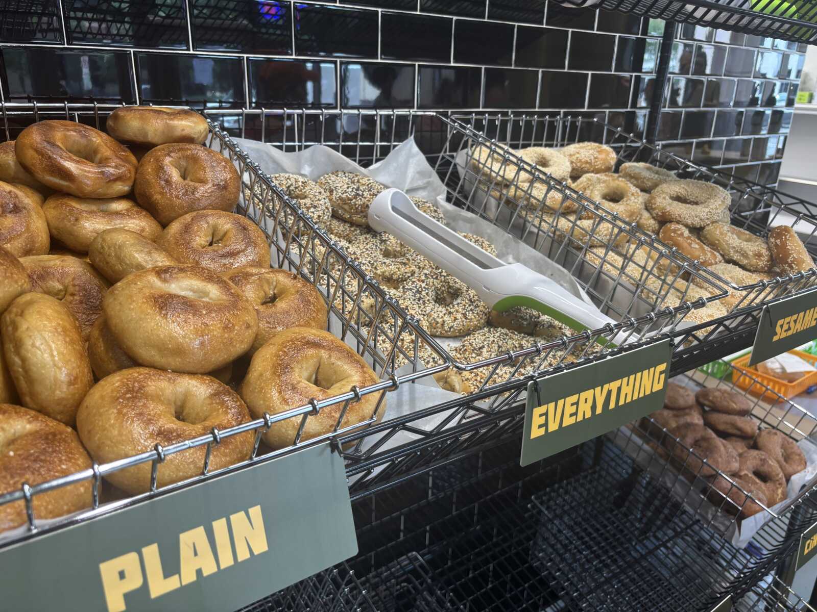 Assorted bagels and donuts in a store display: plain bagels on the left, sesame-covered bagels and donuts in the middle and right; metal racks and tongs nearby.