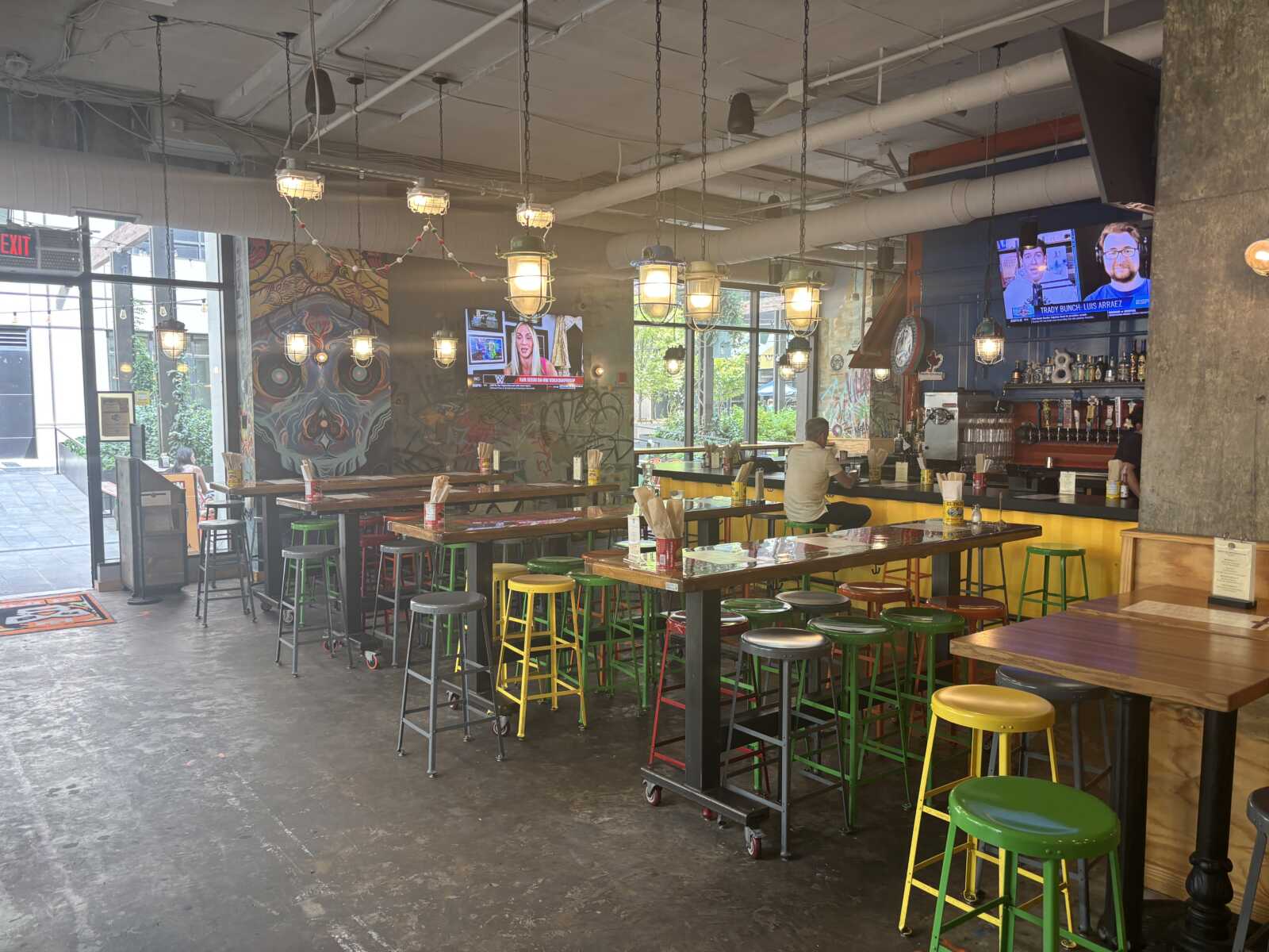 Industrial-style cafe with long communal tables, colorful stools, graffiti wall, and hanging lantern lights.