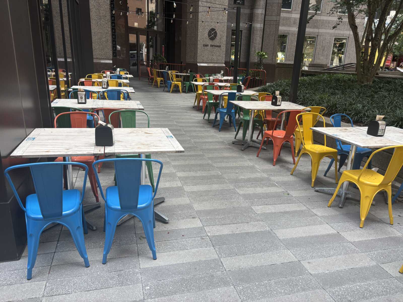 Outdoor cafe seating with colorful metal chairs and white wooden tables along a paved walkway; string lights overhead and modern buildings in the background, mostly empty.
