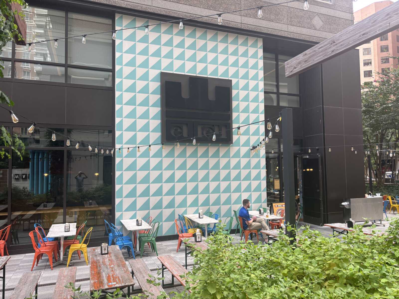 Outdoor cafe seating with colorful metal chairs and weathered wooden tables in front of a turquoise-and-white triangular tiled wall under string lights.