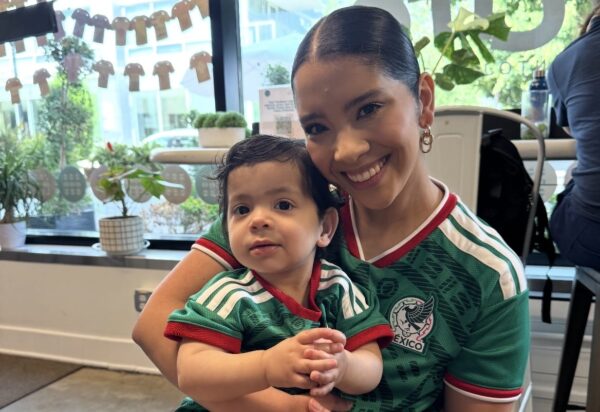Smiling woman with a sleek bun holding a baby in a green Mexico soccer jersey, seated indoors by a window with plants and decorations in the background.