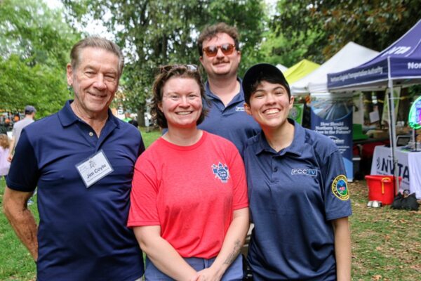 Four people posing for a photo at an outdoor park festival, with vendor tents and green trees in the background.