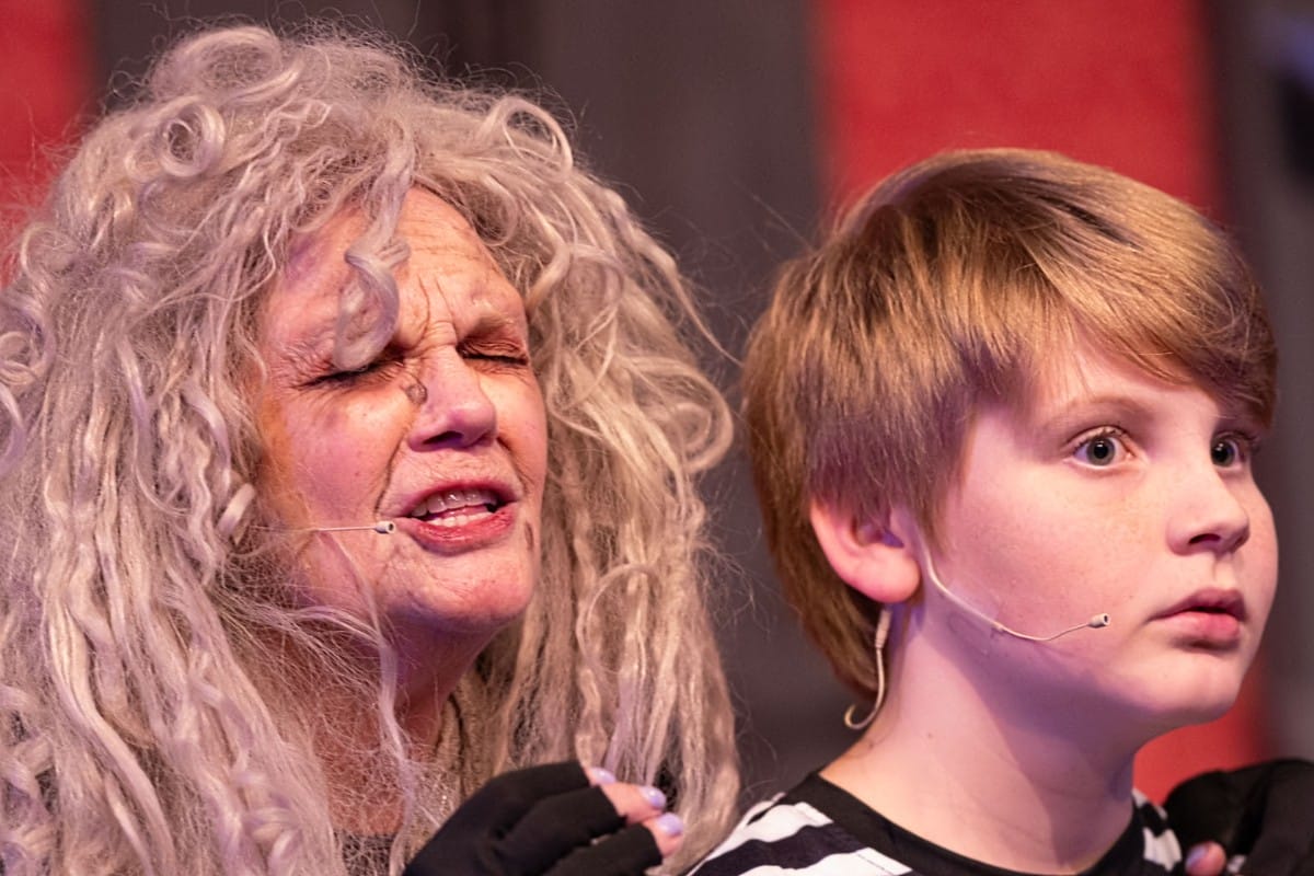 Two performers on stage: an elderly woman with long, curly gray hair and a young boy wearing head-mounted microphones, both focused ahead.