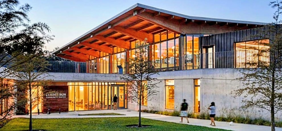 Modern two-story building with large glass walls and a sloped wooden roof at dusk; people on the walkway.