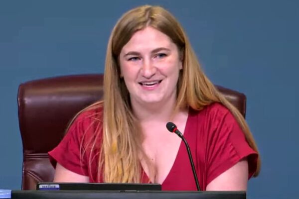 Woman in a red blouse sits at a desk with a microphone, speaking at a formal meeting.