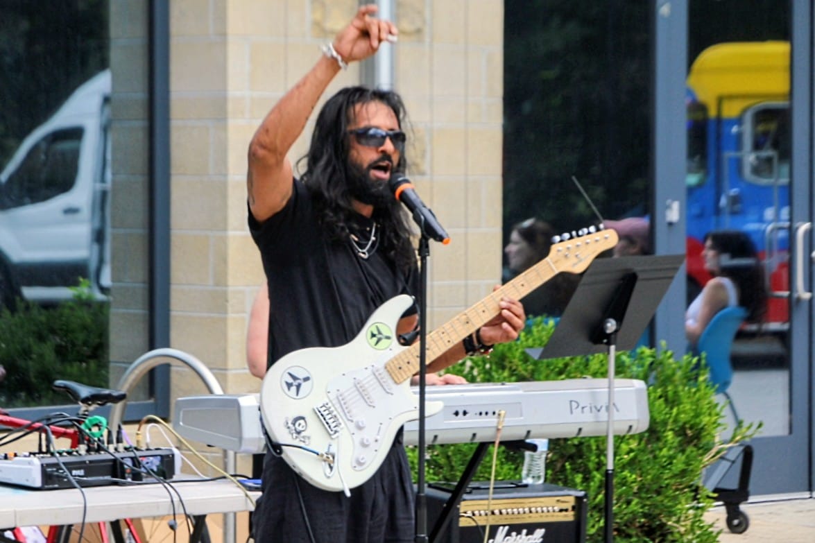Male guitarist with long hair and sunglasses sings into a microphone while playing a white electric guitar outdoors.