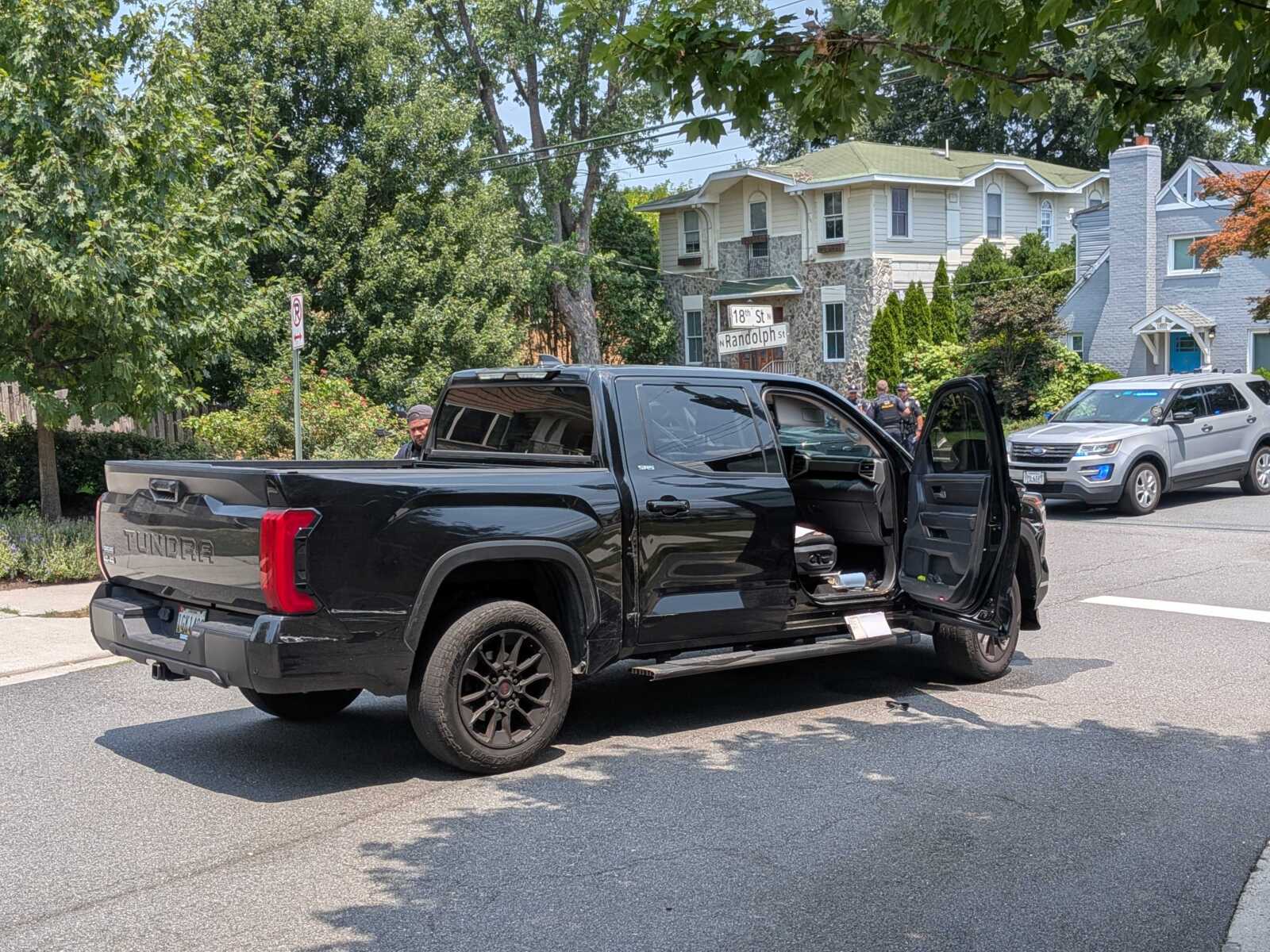 Black pickup truck with its doors open parked on a residential street, with officers and a police SUV in the background among trees and houses.