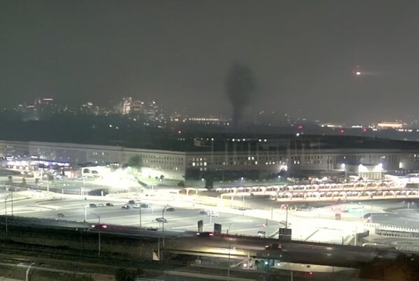 Nighttime cityscape with a dark plume rising from far buildings, seen beyond a lit rail yard and parking area.
