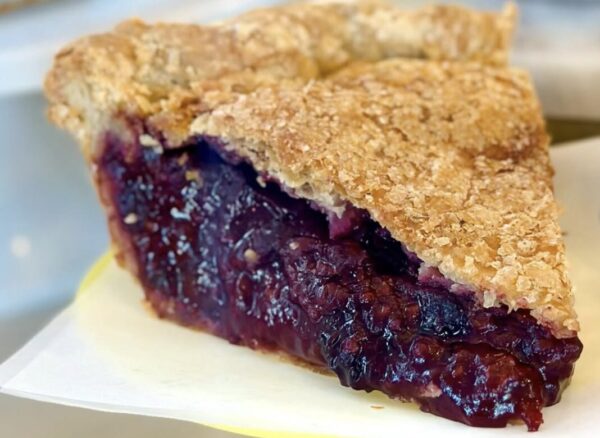 Slice of crumb-topped blueberry pie on a plate, showing a glossy purple filling and golden crust crumb.