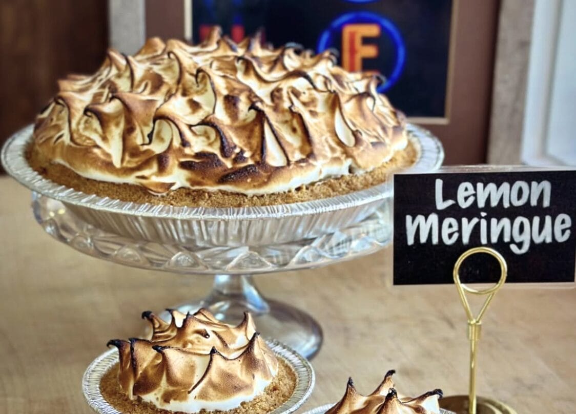 Two lemon meringue pies with toasted peaks on a glass cake stand, a sign reads 'Lemon meringue' nearby.