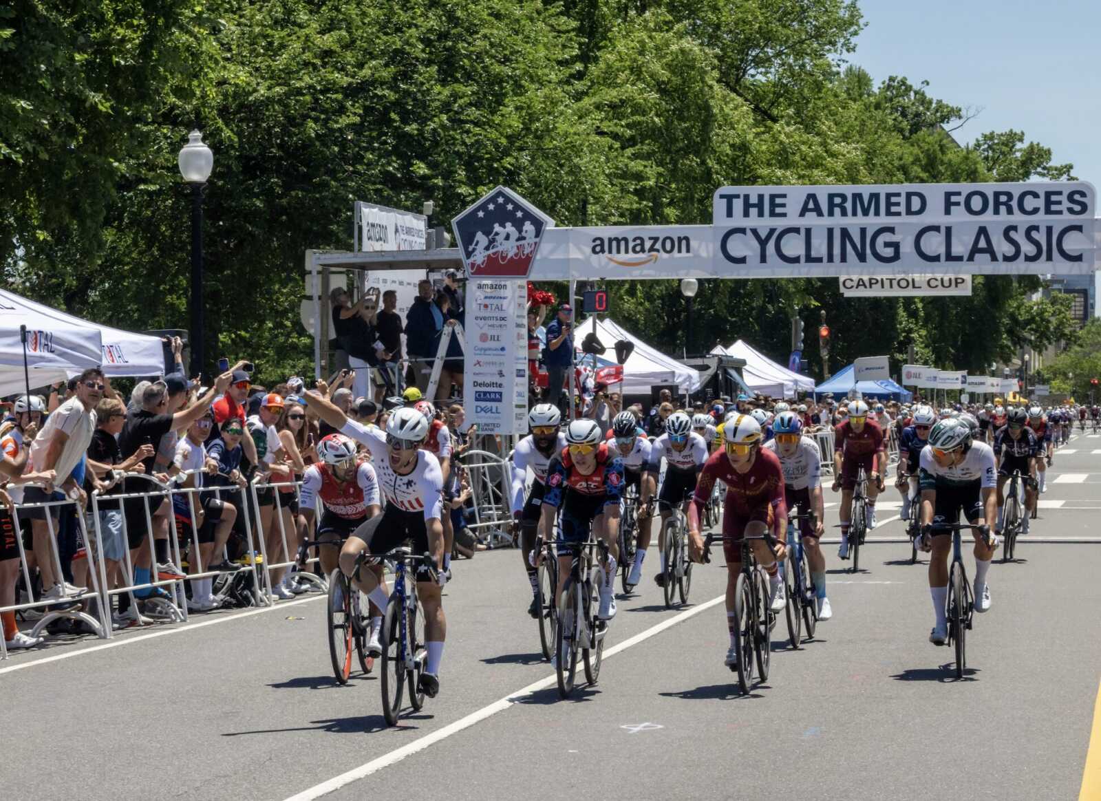 Cyclists sprint from the start under a banner reading The Armed Forces Cycling Classic, with cheering spectators along barricades and tents on the side street.
