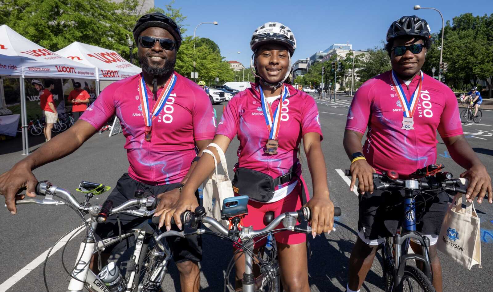 Three cyclists in pink jerseys with helmets and medals pose with their bikes on a city street after a race, event tents in the background.