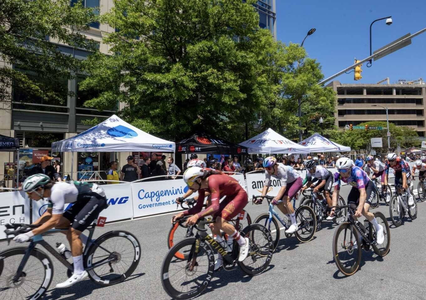 A group of cyclists race down a city street lined with white tents and barriers, with spectators watching from behind the barriers.