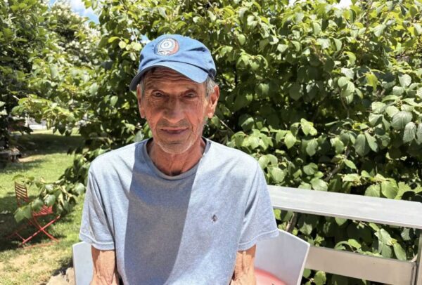 Older man wearing a blue cap and gray T-shirt sits on a bench outdoors with lush green foliage behind him.