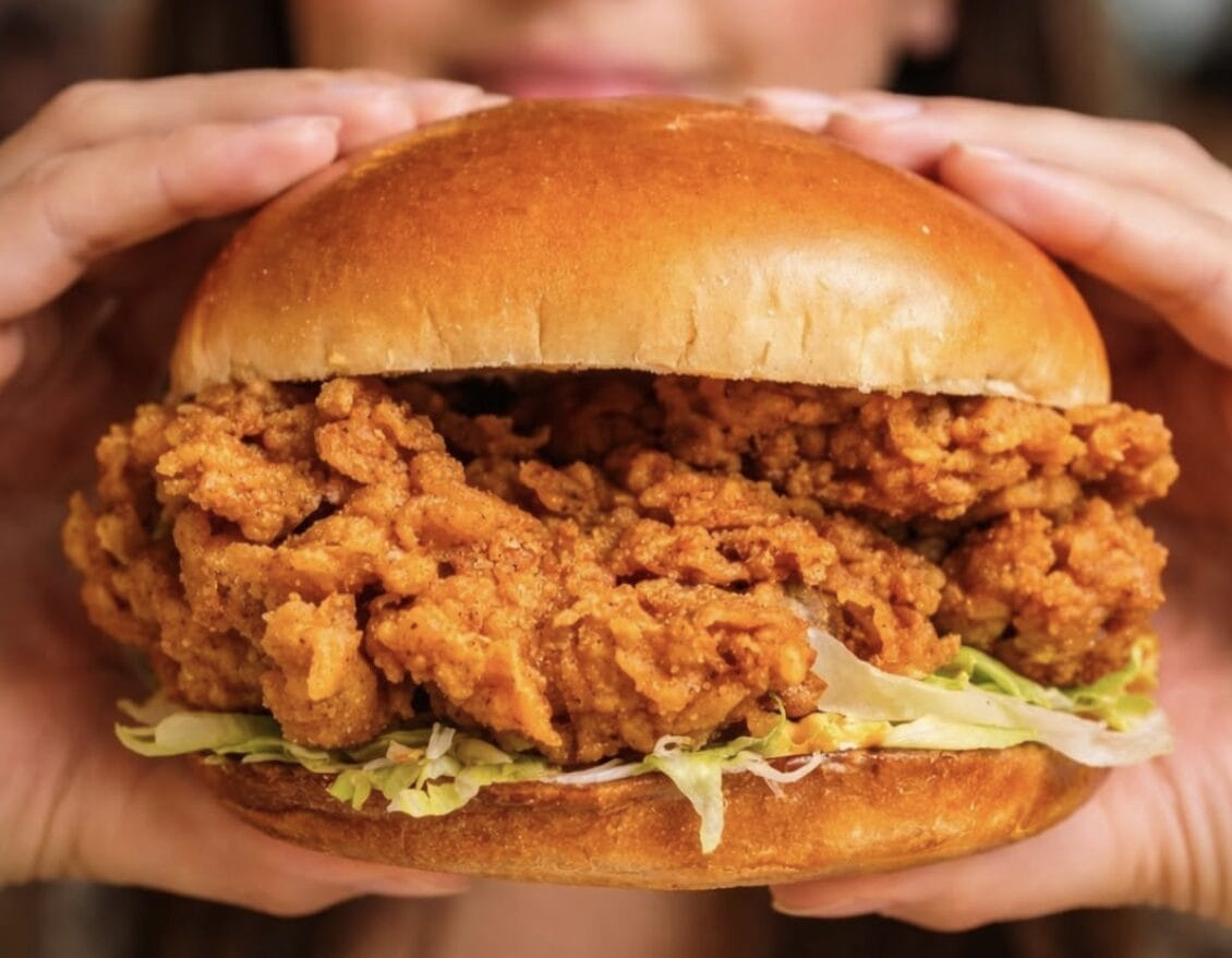 Hands hold a large crispy fried chicken sandwich with shredded lettuce on a soft bun.