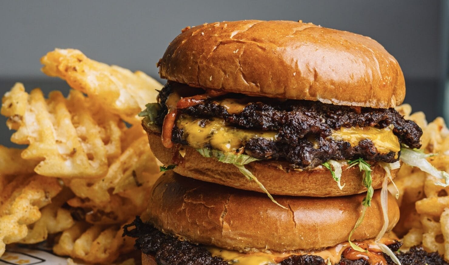 Two-patty cheeseburger with melted cheddar, lettuce, onions on a glossy brioche bun, paired with waffle fries in the background.