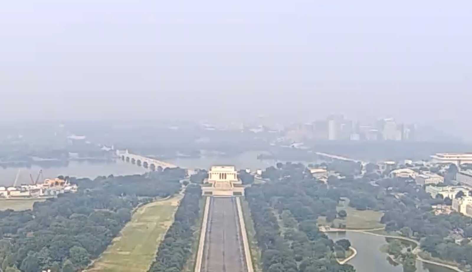 Aerial view of a long central avenue in a urban park, lined with trees, leading to a large beige monument, with a river and distant city skyline in the background.