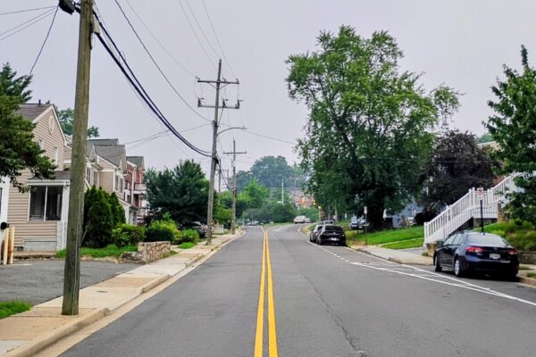 Residential street with parked cars, trees, and utility poles along a two-lane road to the horizon.