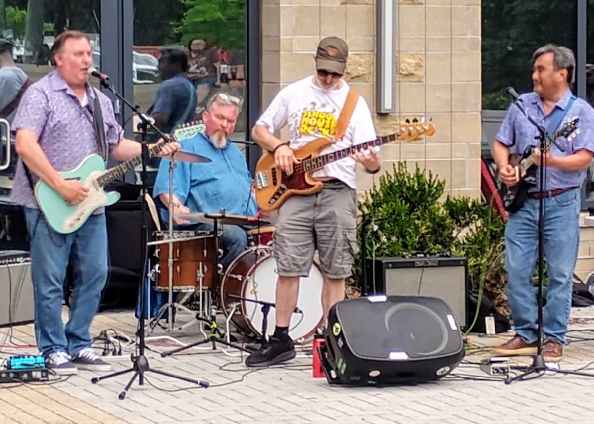 Four musicians play outdoors: left guitarist with mint-green guitar, drummer in blue, center-left bassist wearing a cap, right guitarist with dark guitar.