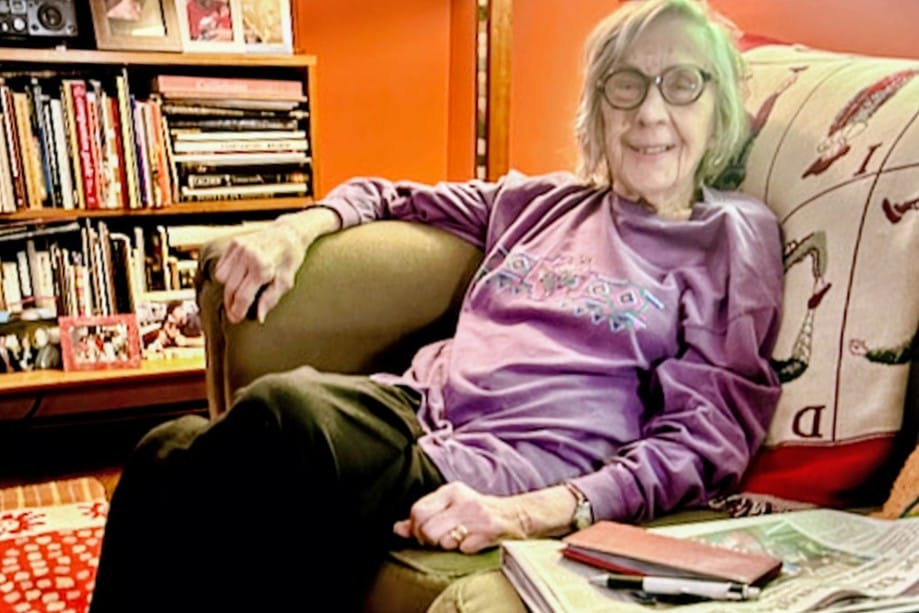 Vivian Kallen in later years (via Arlington AAUW) | ARLnow Older woman with glasses sitting on a brown sofa in a cozy living room, wearing a purple sweatshirt, smiling at the camera with a bookshelf behind her.