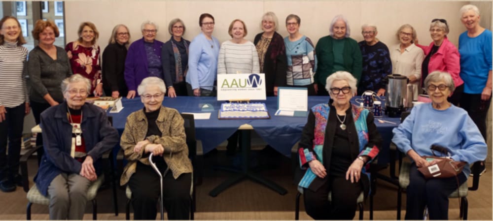 Vivian Kallen, seated at right, at Arlington AAUW event (via Arlington AAUW) | ARLnow Group of about 20 seniors gathered in a room for a celebration, with a cake and AAUW sign on the table at center. Two rows: several people seated in front and others standing behind them.