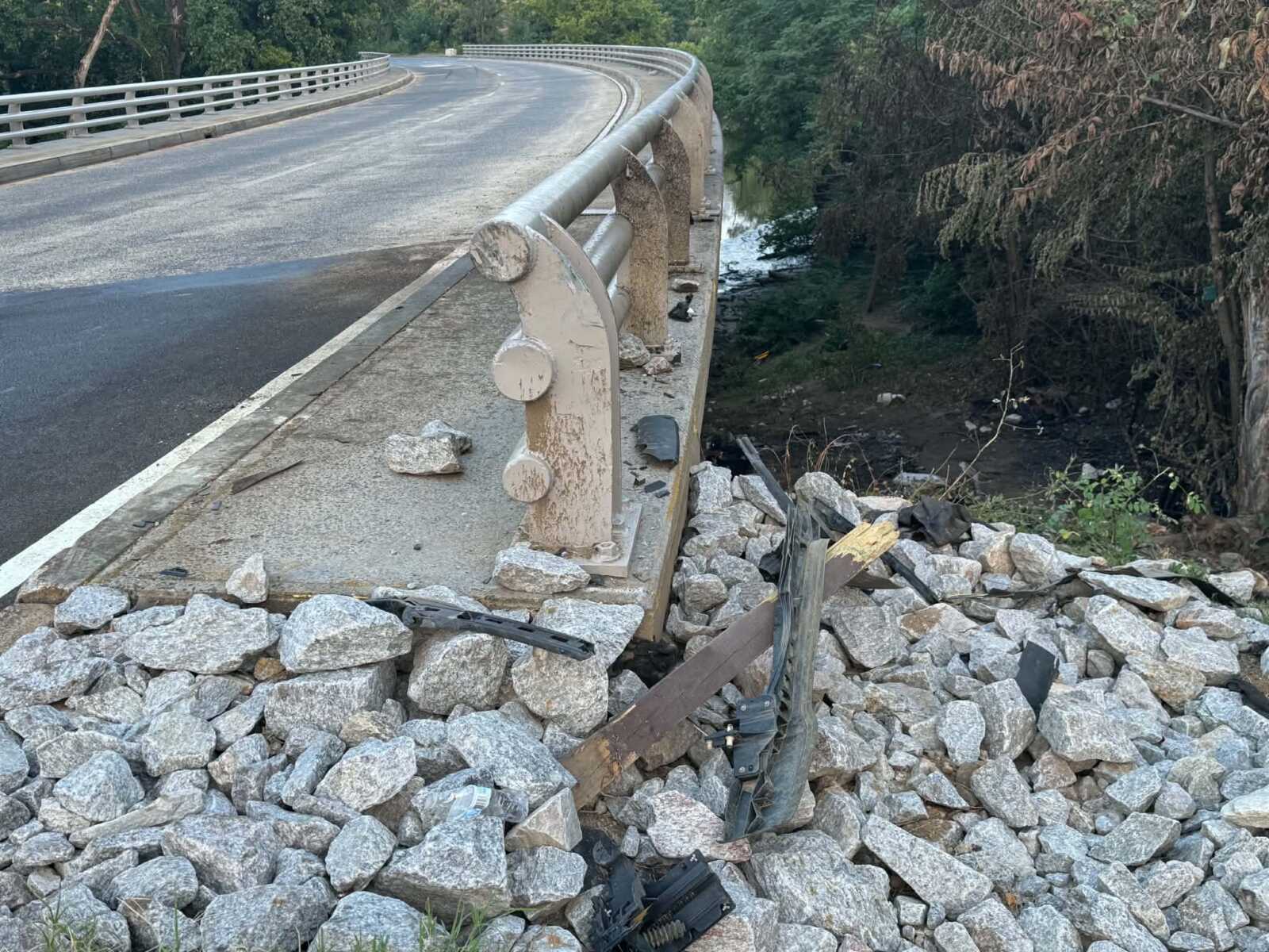 Curved bridge with concrete guard rails over a rocky drop; debris and rubble along the edge with trees beyond the edge of the road.