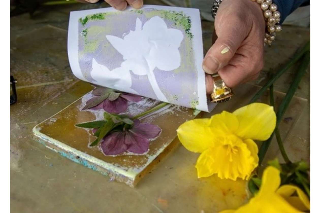 Close-up of hands pressing a flower-printed sheet onto a surface beside purple flowers and bright yellow daffodils.