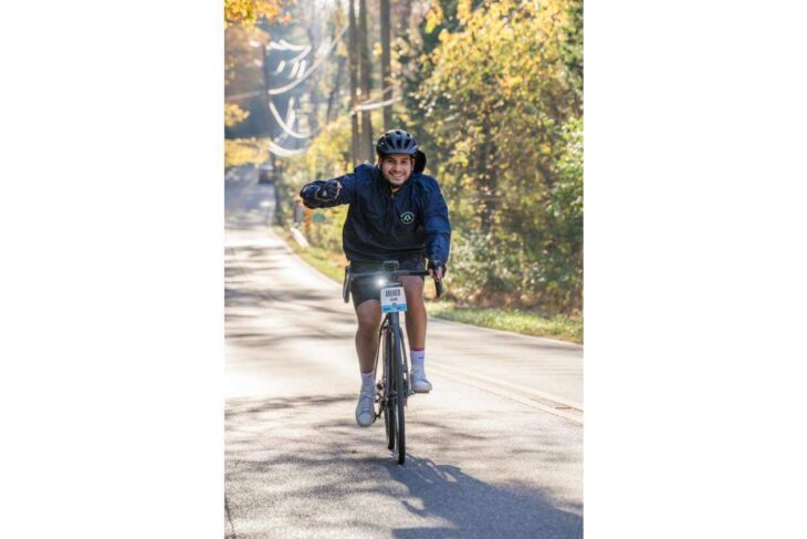 Man wearing a helmet rides a road bike toward the camera on a sunlit autumn path, giving a thumbs-up.