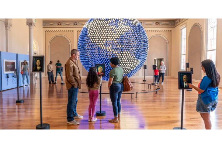 Visitors in a museum view portrait kiosks around a large blue-and-white dotted sphere sculpture centerpiece.