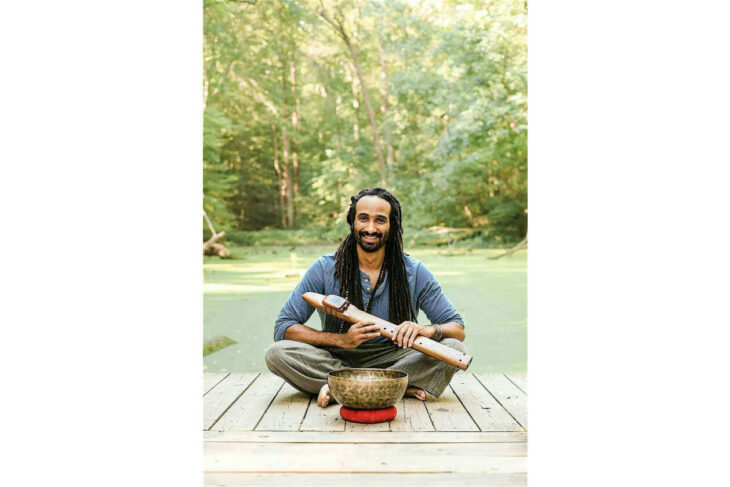 Smiling man with dreadlocks sits cross‑legged on a wooden dock by a pond, holding a wooden flute with a singing bowl in front.