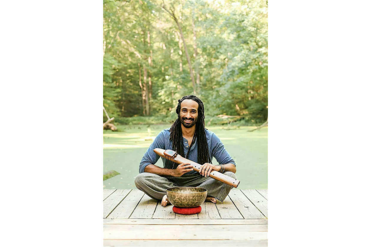 Smiling man with dreadlocks sits cross‑legged on a wooden dock by a pond, holding a wooden flute with a singing bowl in front.
