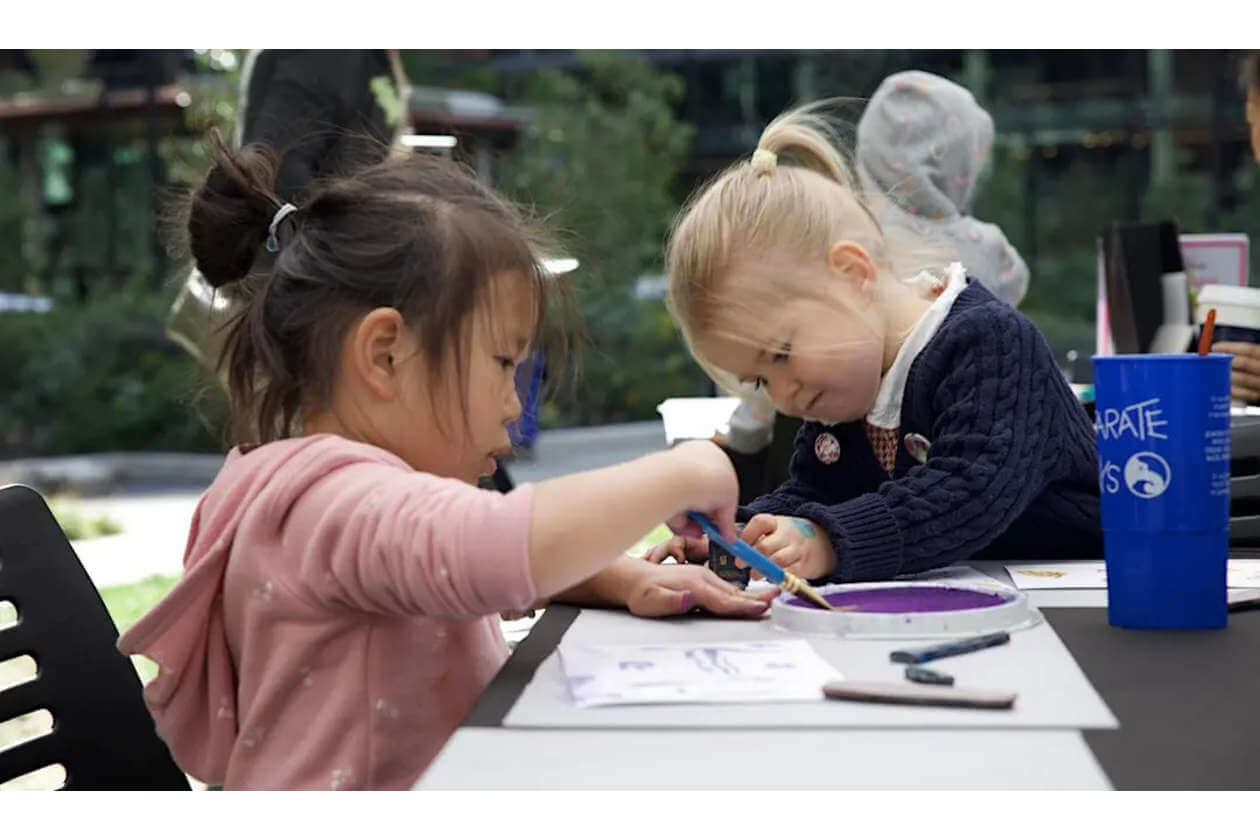 Two young girls sit at a table outdoors, focused on painting with brushes and purple paint on paper.