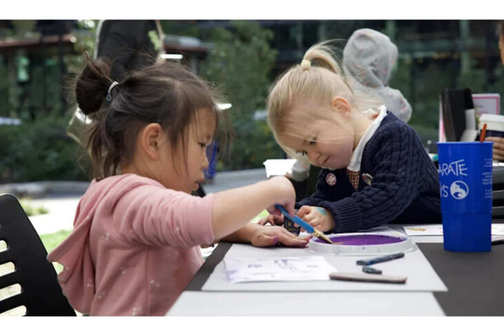 Two young girls sit at a table outdoors, painting with brushes and purple paint, focused on their art project.