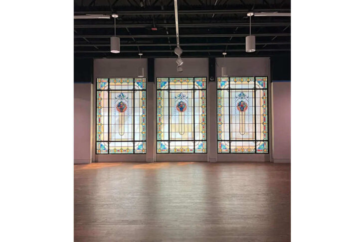 Three tall decorative stained-glass panels with blue, orange, and yellow motifs in a modern gallery space under track lighting.