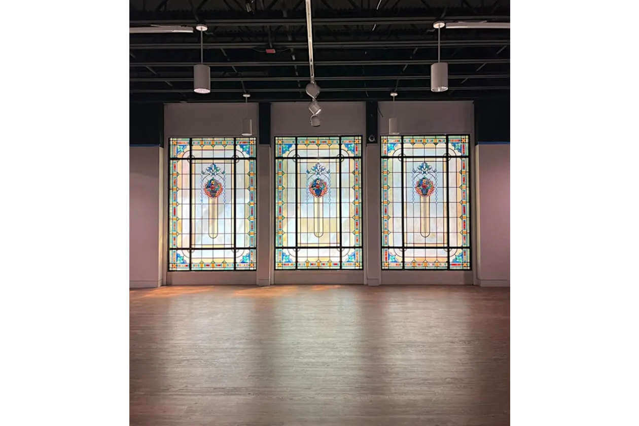 Three tall, colorful stained-glass windows in an empty gallery with overhead track lighting and a wooden floor.