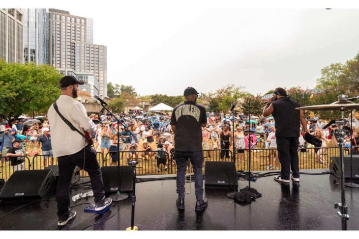 Three musicians perform on an outdoor stage facing a large crowd, with city buildings and trees in the background.