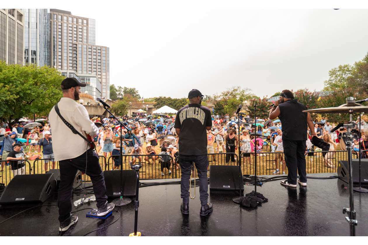 Three musicians perform on an outdoor stage facing a large crowd, with city buildings and trees in the background.