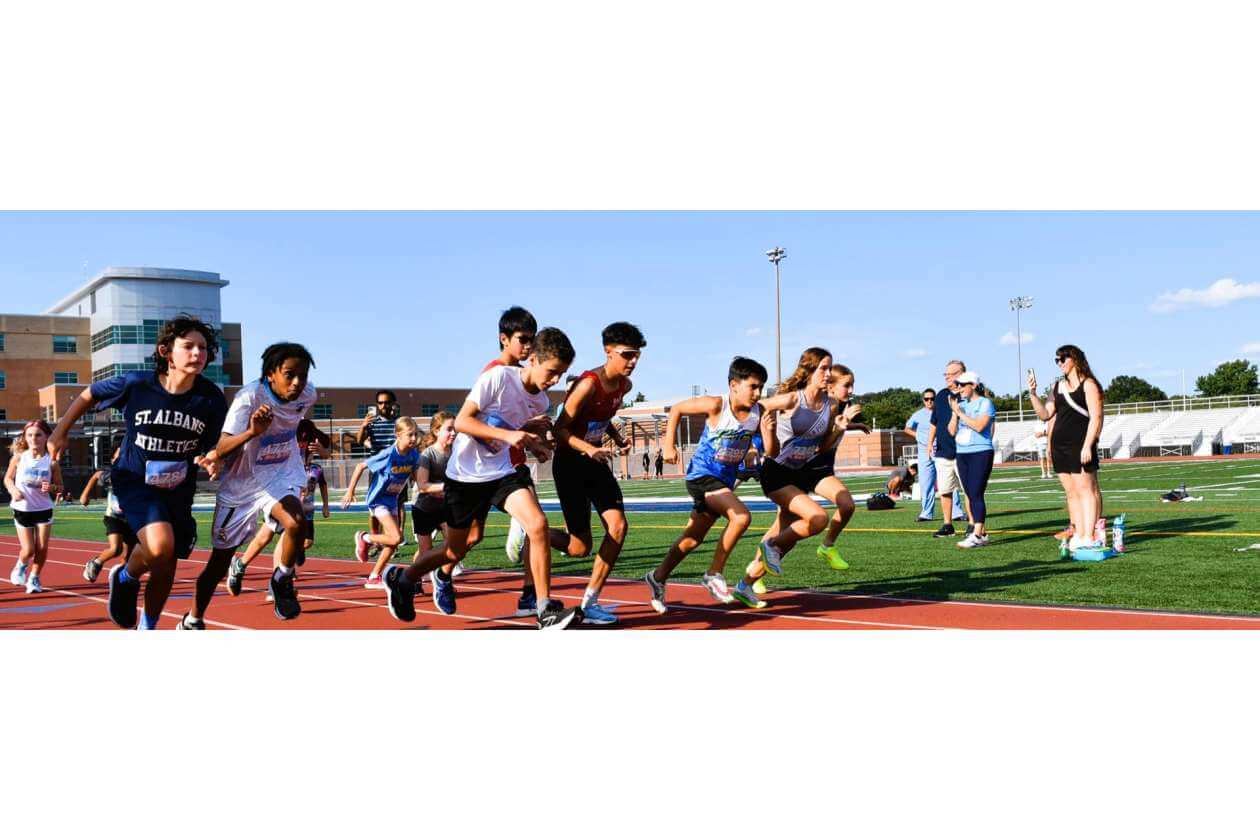 Group of children sprinting at the start of a race on an outdoor track, with a stadium and spectators in the background