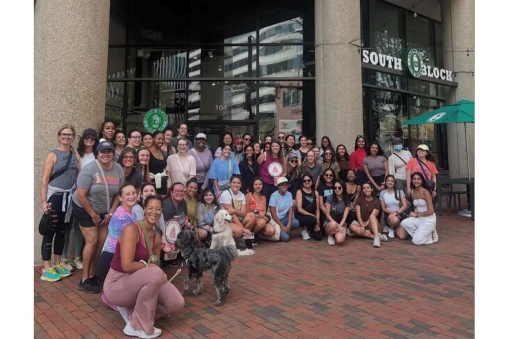 Large group of women posing together outside a building with a South Block sign in the background; two dogs sit at the front on a brick-paved area under a green umbrella nearby.