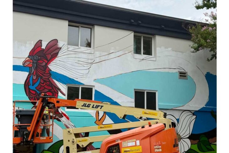 Large mural of a red rooster painted on the side of a building, with an orange boom lift in the foreground.