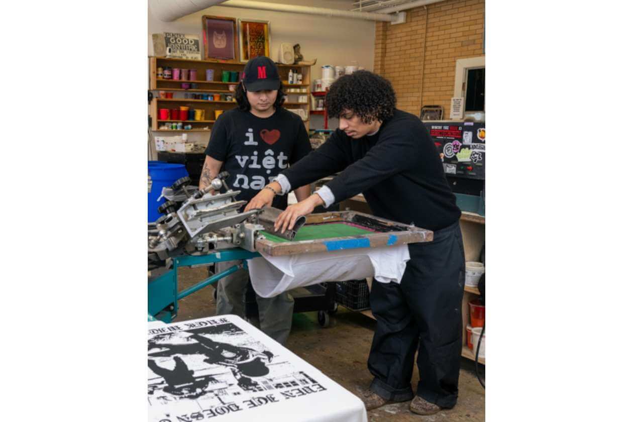 Two people operate a screen-printing press, transferring ink to a white T-shirt in a cluttered workshop with shelves in the background.