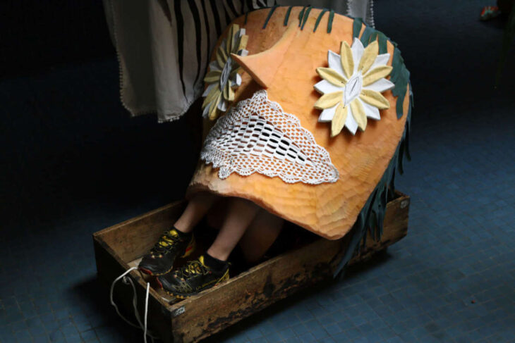 Person seated in a wooden sled wearing an orange quilted cape with lace trim and large flower appliqués.