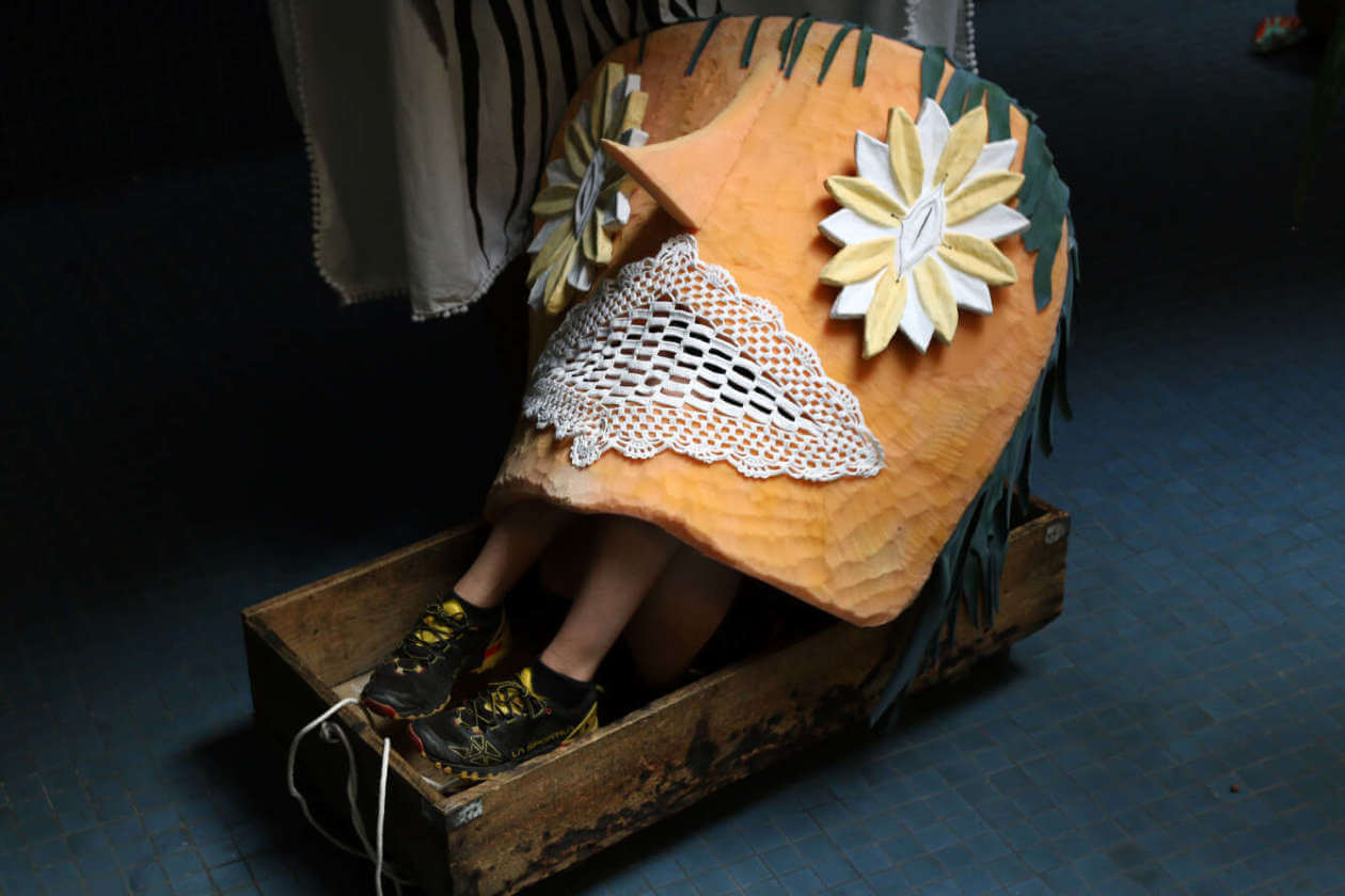 Person seated in a wooden sled wearing an orange quilted cape with lace trim and large flower appliqués.