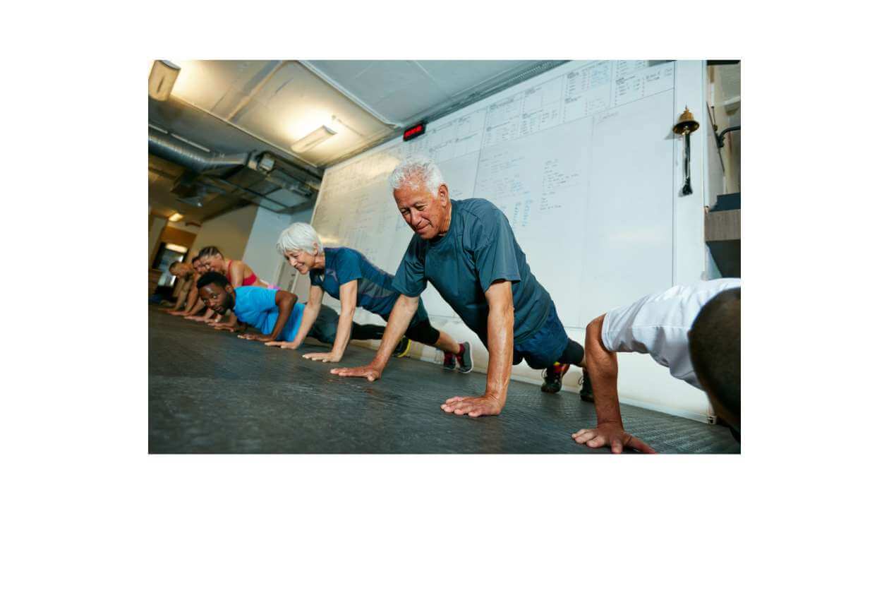 Group of people in a gym doing push-ups on a mats-lined floor, led by an older man in a blue shirt next to a whiteboard.