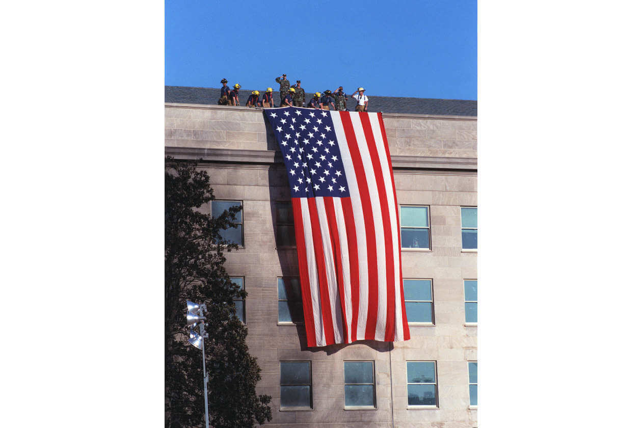 Large American flag hanging down the side of a beige stone building, with people standing on the roof under a clear blue sky