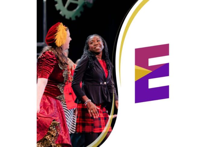 Dancers performing on a stage beside a large purple E logo on a white backdrop; a smiling woman in a red plaid skirt is in the foreground.